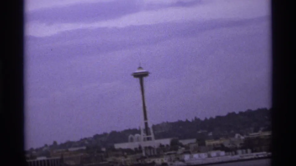 1970s 8mm frame showing the Space Needle from across water, with low buildings and wooded hills below a muted sky.