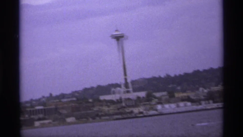Handheld 8mm frame with the Space Needle slightly off center, water in the foreground, and shoreline buildings below wooded hills.