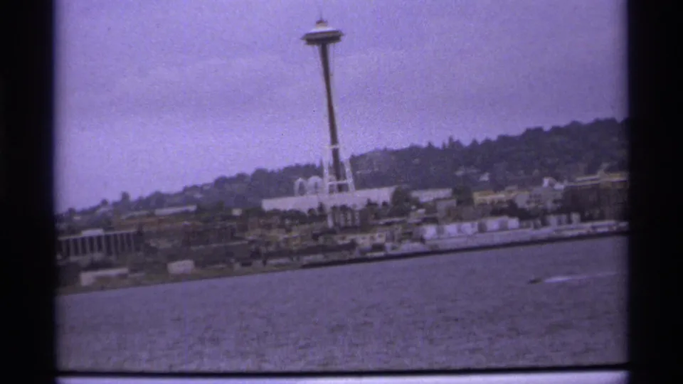 1970s 8mm frame appears to show the Space Needle across water, with shoreline buildings and a wooded hillside behind it.