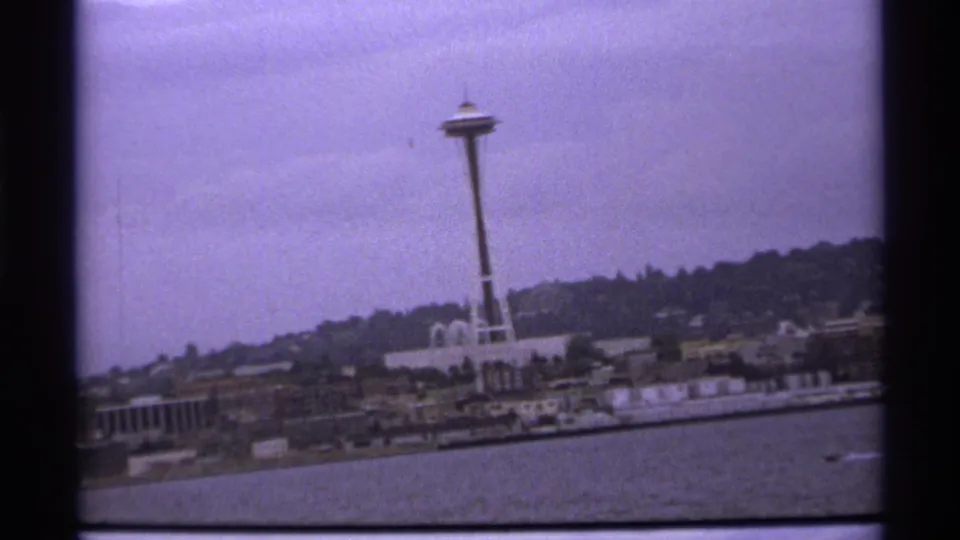 The Space Needle stands above a waterfront district, with open water in the foreground and a faint boat wake to the right.