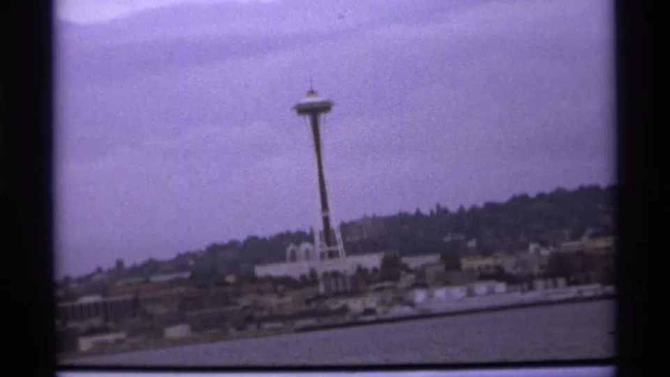 A handheld 8mm frame shows the Space Needle above shoreline buildings, with water below and dark hillside behind.