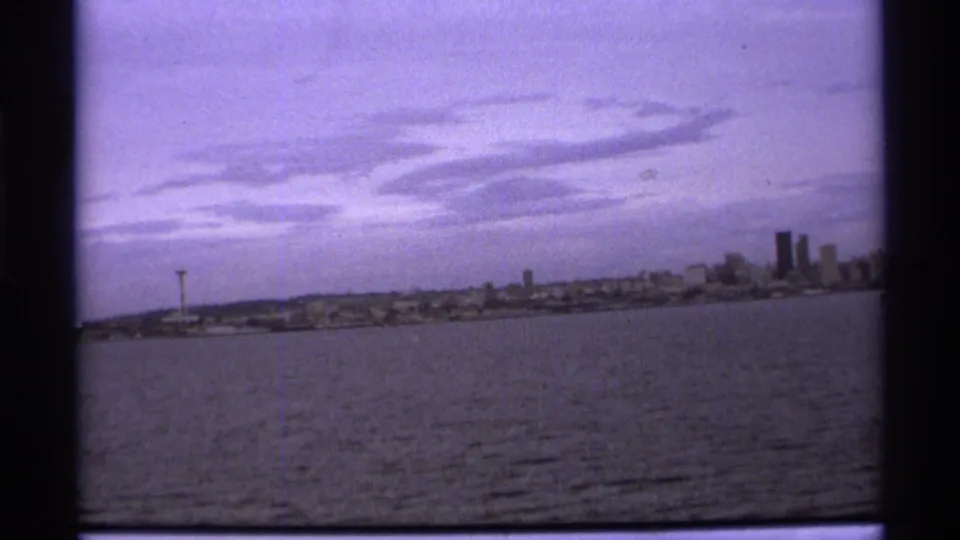 1974 8mm view of a distant Vancouver skyline across water, with a needle-like tower at left and high-rises at right.