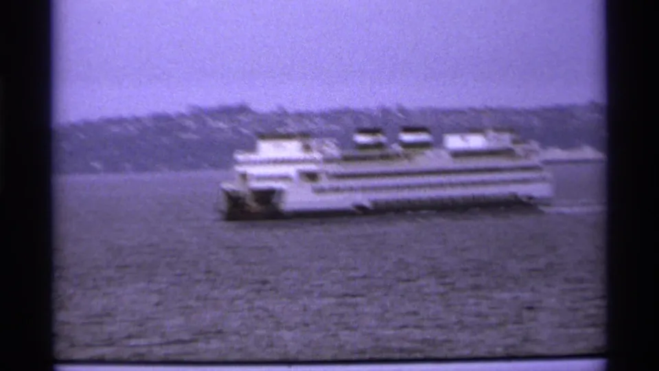 1974 8mm frame of a large white ferry moving across the water, with a hazy shoreline and hills in the background.