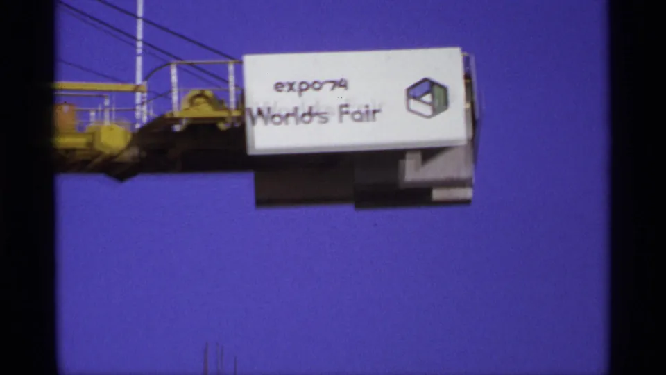 Soft handheld 8mm frame of the Expo '74 World's Fair sign suspended from a yellow crane arm against a saturated blue sky.