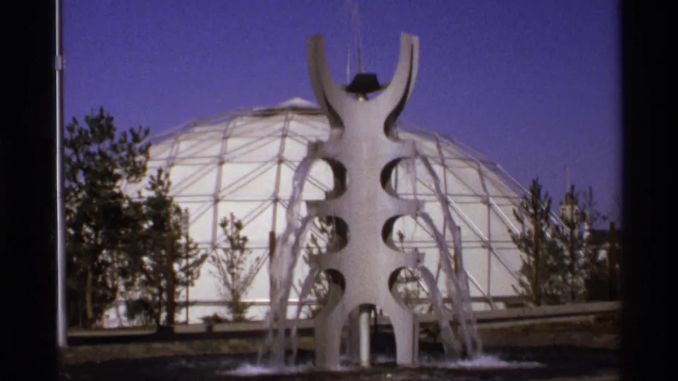 8mm frame showing a branching sculptural fountain spraying water before a large geodesic dome, with trees along the base.