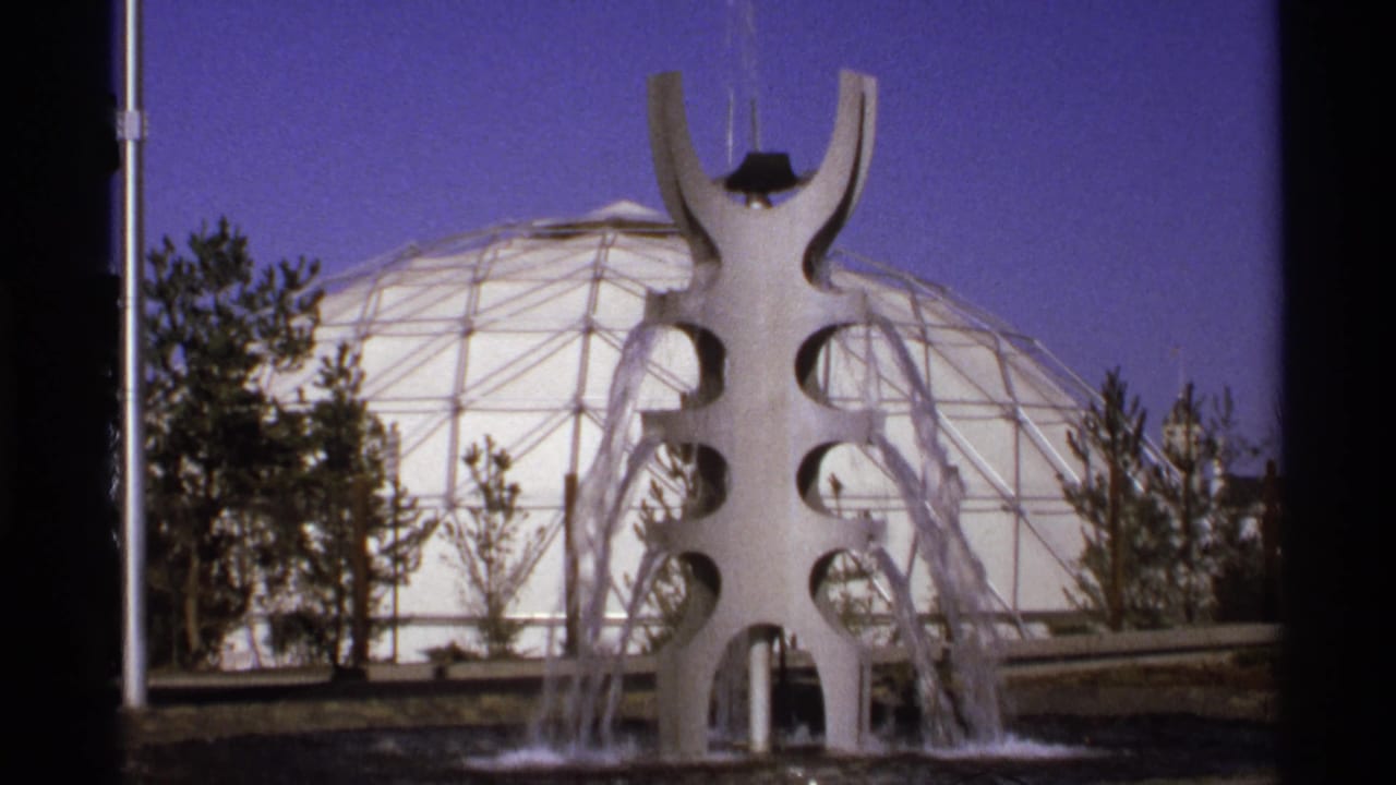 1970s 8mm frame of a white public fountain with water jets before a large geodesic dome, with trees and blue sky behind it.