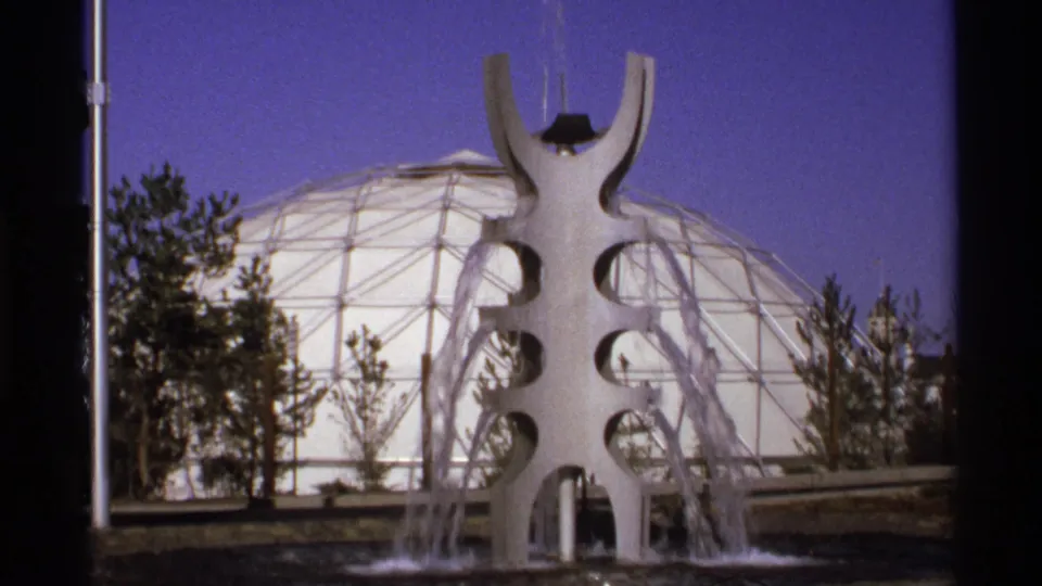 1970s 8mm frame of a white public fountain with water jets before a large geodesic dome, with trees and blue sky behind it.