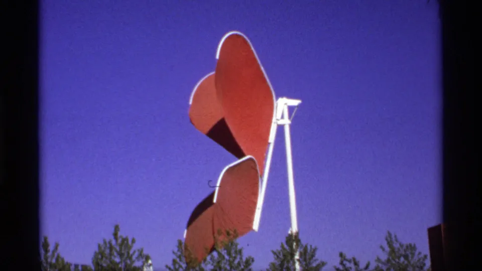 1970s 8mm frame of a red abstract butterfly-like sculpture mounted on a white support, seen above trees against a deep blue sky.
