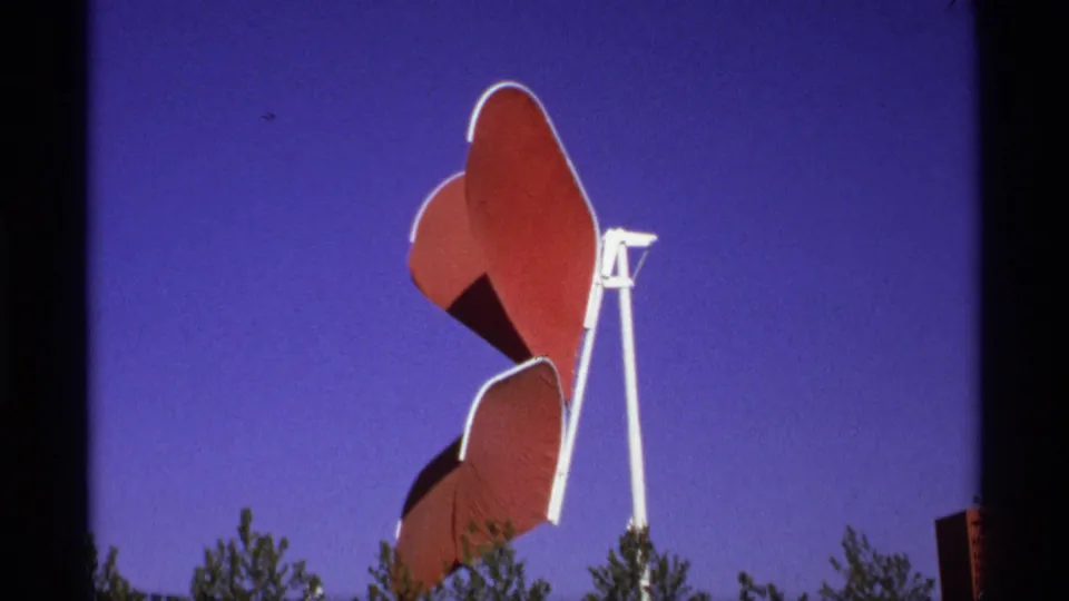 Close visitor-shot view of a red butterfly-like public sculpture on a white frame, with tree tops low in the 8mm frame.