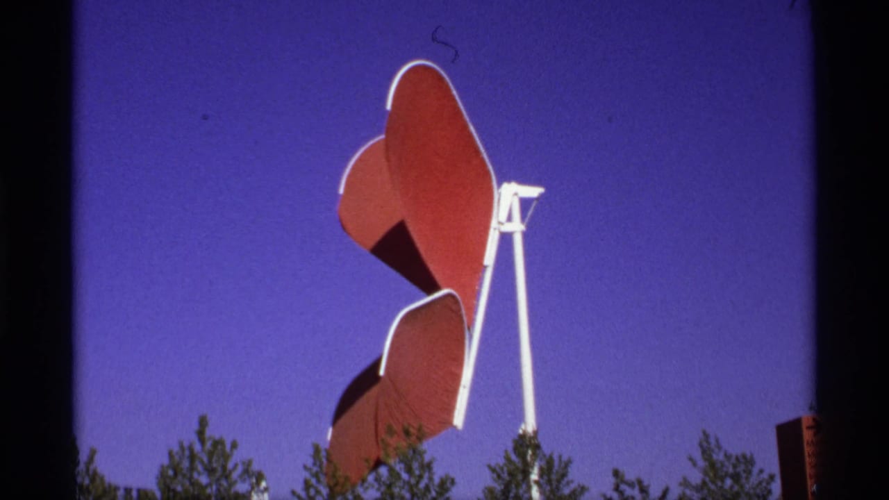 Red and white outdoor sculpture above treetops against a blue sky.