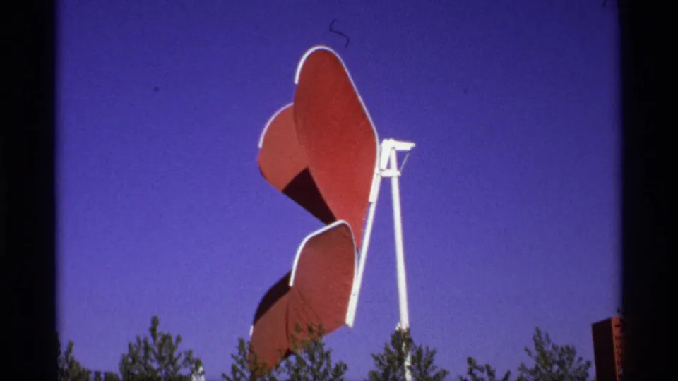 1970s 8mm frame of a red and white sculptural installation rising above treetops against a saturated blue sky.