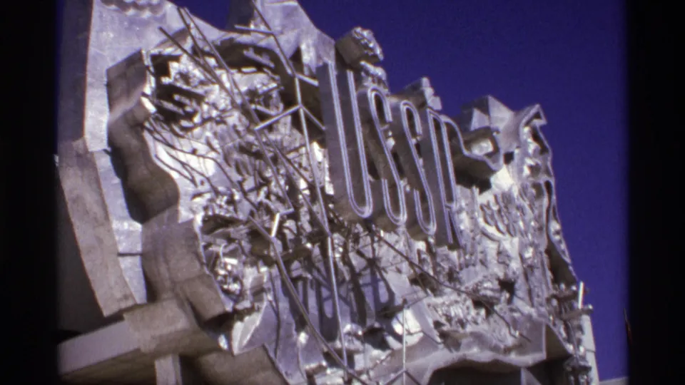 8mm close-up of the USSR display sign, showing raised letters, silver relief forms, and sky in the background.