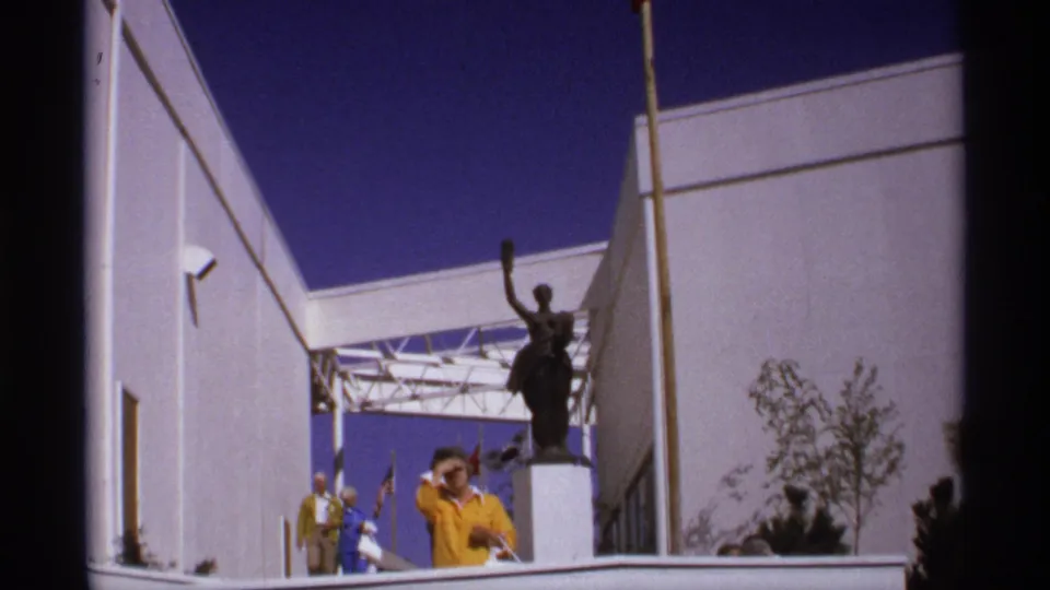 Visitors at the World's Fair walk toward the camera in a courtyard beside a raised-arm statue and modern building walls.