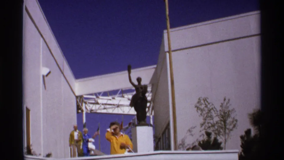 End frame showing visitors walking through a fair courtyard, with a dark statue, pale walls, shrubs, and blue sky behind them.