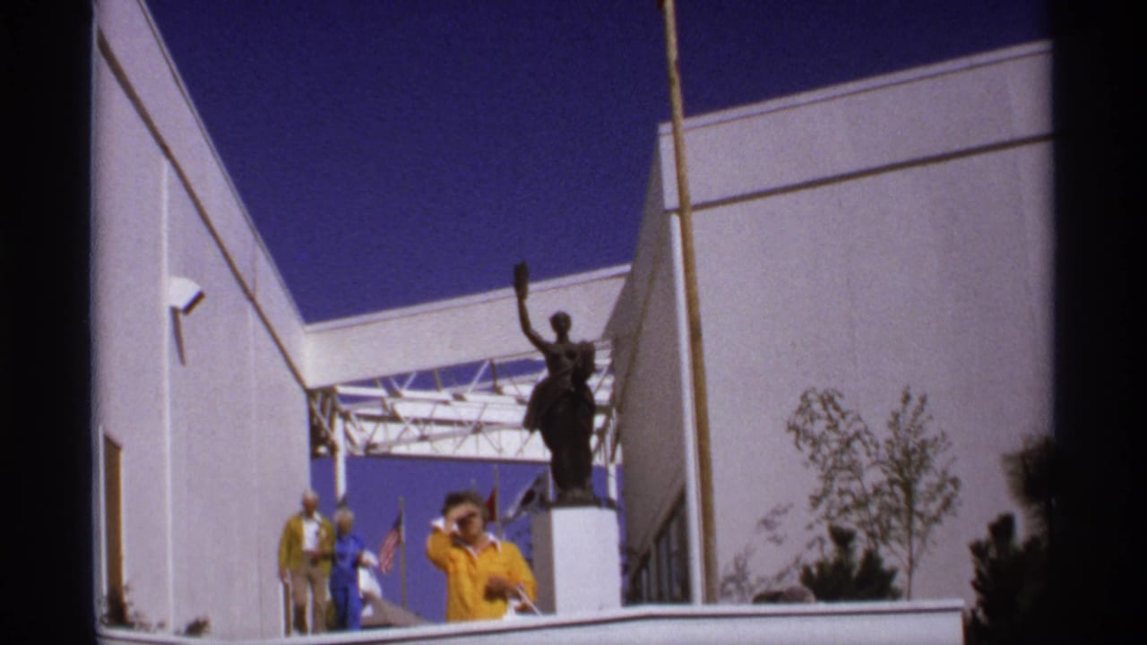 1974 8mm view of a raised-arm statue outside a white fair building, with visitors standing nearby under a deep blue sky.