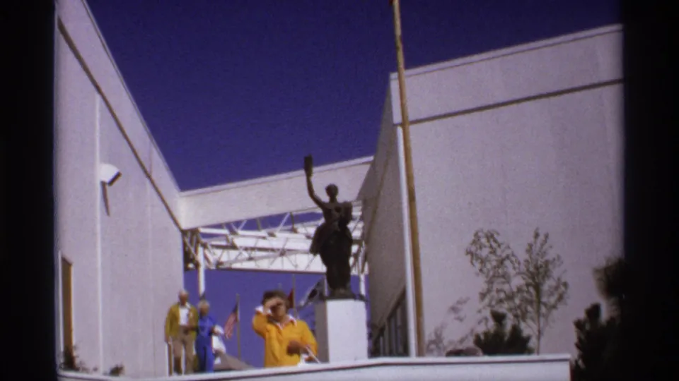 1974 8mm view of a raised-arm statue outside a white fair building, with visitors standing nearby under a deep blue sky.