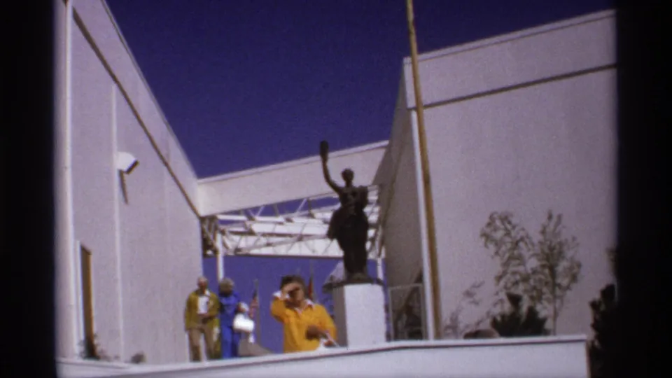 1974 fairground exterior with a raised-arm statue, visitors in period clothing, and white modern pavilion architecture.