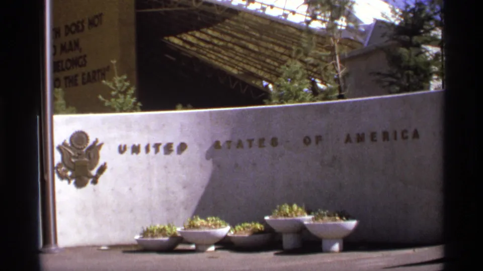 Close 1974 8mm view of a concrete wall marked United States of America, with U.S. seal, planters, and partial pavilion structure above.