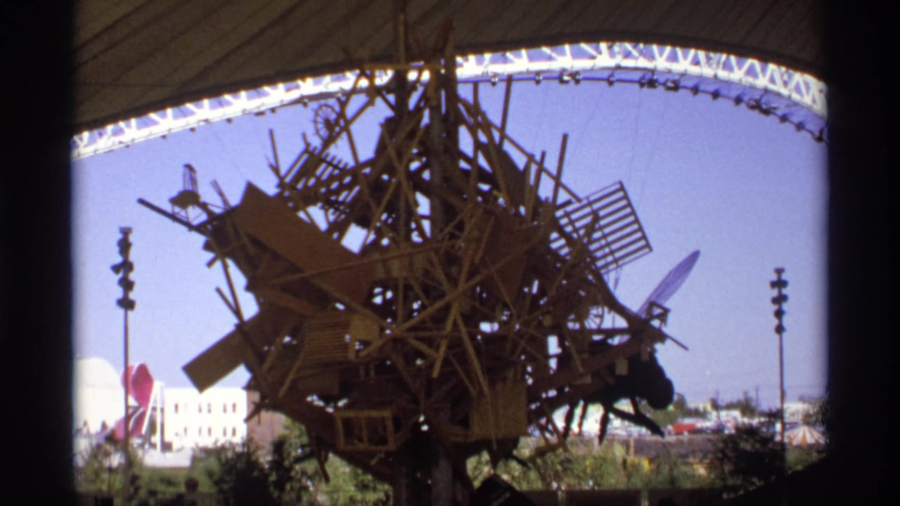 Large wooden assemblage sculpture under a white arched canopy with fairground structures behind it.