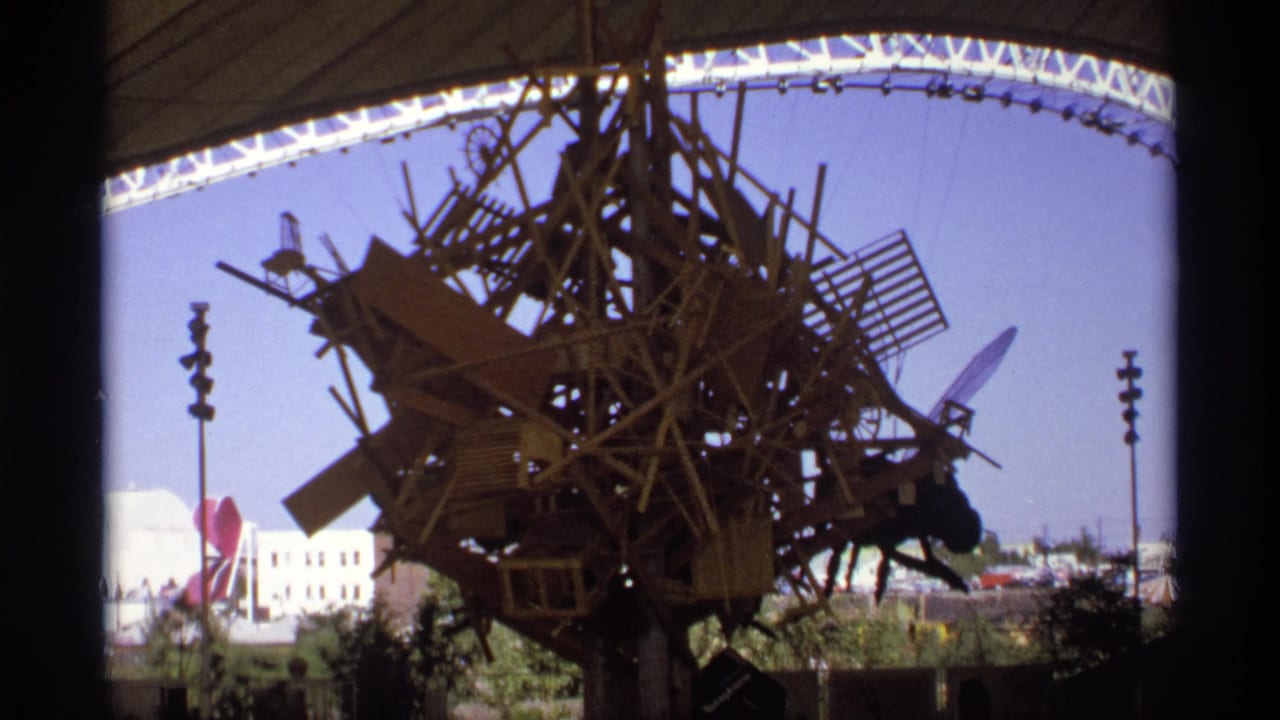 A visitor-shot 8mm frame shows a tall assemblage of wood and debris beneath a broad white canopy, with open fairground space in the background.