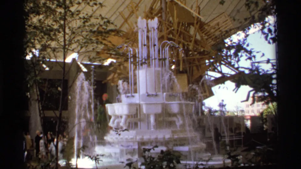 A multi-tiered white fountain made from sinks and vertical pipes pours water in a landscaped area beneath a canopy, with visitors partly visible nearby.