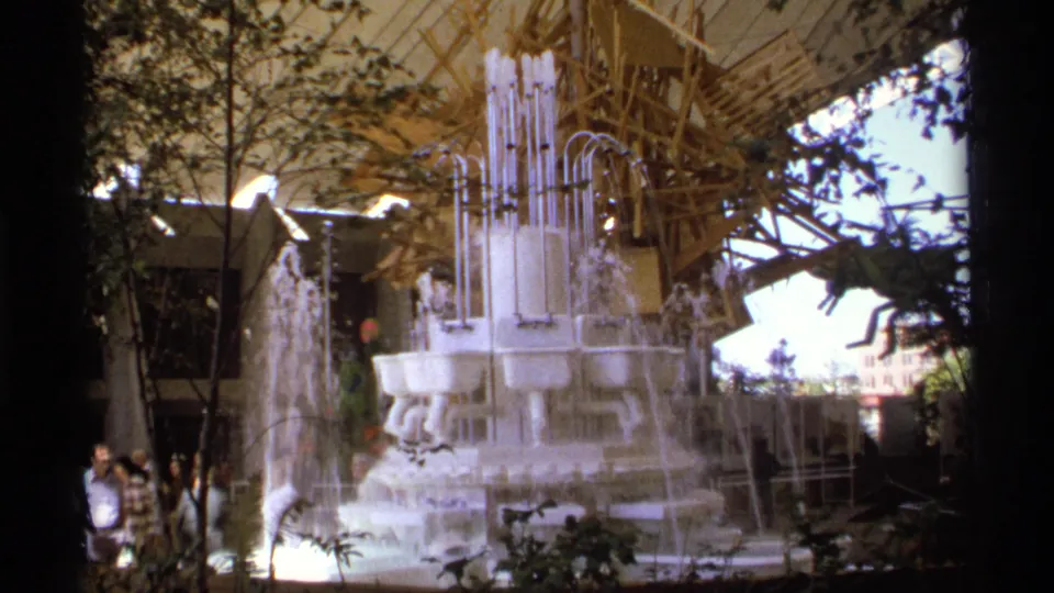The 8mm frame lingers on the white sink fountain as water falls through several levels, with trees, canopy supports, and visitors at the edge of the scene.