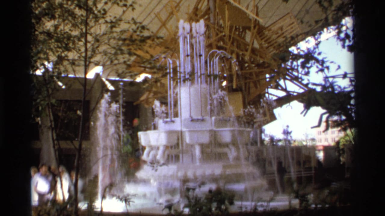 Handheld 8mm view of a bright fountain beneath a tented public pavilion, with visitors standing nearby and dark film borders visible.