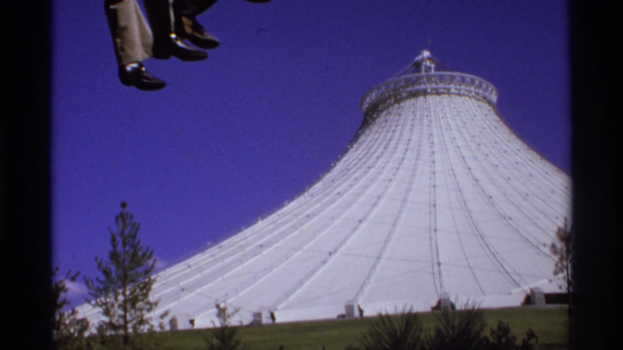 Dangling trouser legs and dress shoes above a white domed roof under blue sky.