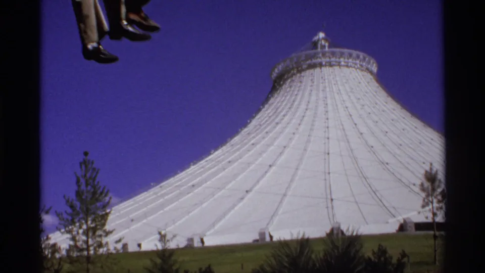 1974 8mm frame of the white Seattle dome identified by metadata, seen from a moving ride with feet entering the upper frame.