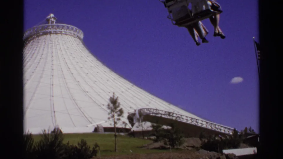 1974 home-movie frame with the white pavilion cropped close, a suspended aerial car above, and a low ramp structure at right.