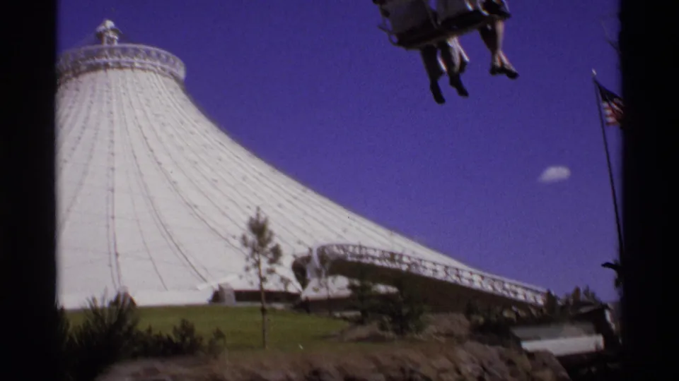White fair pavilion seen from below with suspended riders overhead, a small tree, low entrance structure, and partial flag at right.