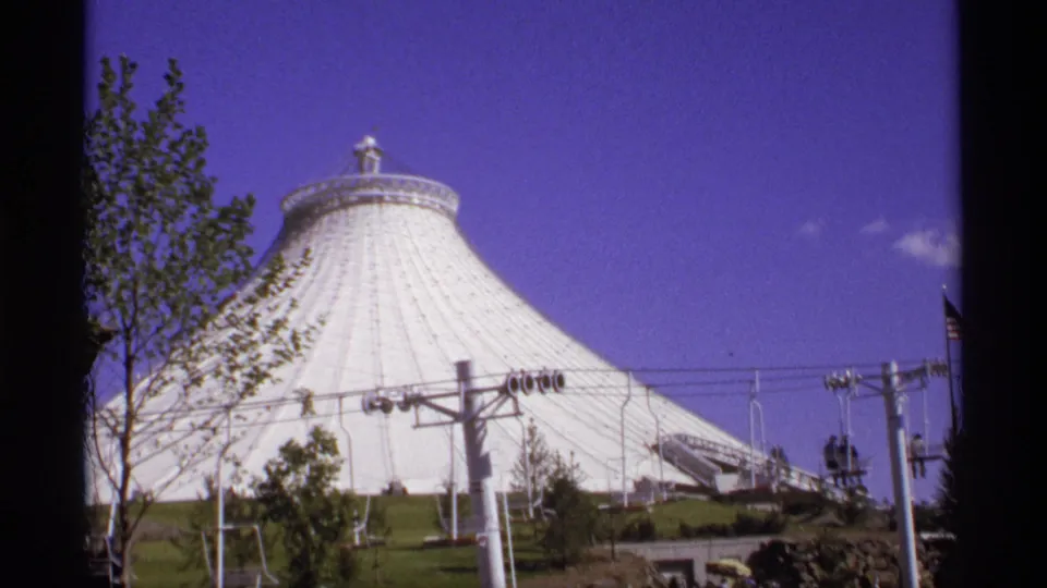 Home-movie frame showing a large white tent-like fairground structure with trees, utility poles, and blue sky.