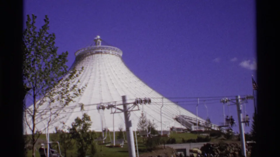 1970s 8mm frame showing a white conical fairground dome, gondola cables, trees, and landscaped grounds under a saturated sky.