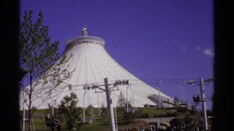 White fairground dome fills the frame behind cable-car towers, small gondolas, trees, and open landscaped space.