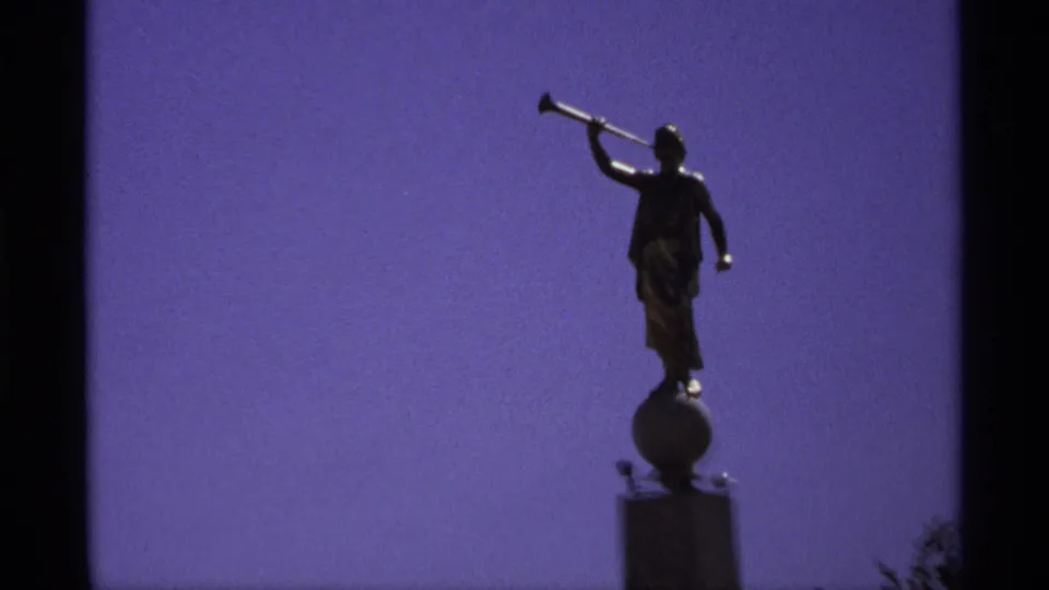 Dark silhouette of a trumpet-bearing statue on a pedestal, isolated against the saturated sky in 8mm home-movie footage.