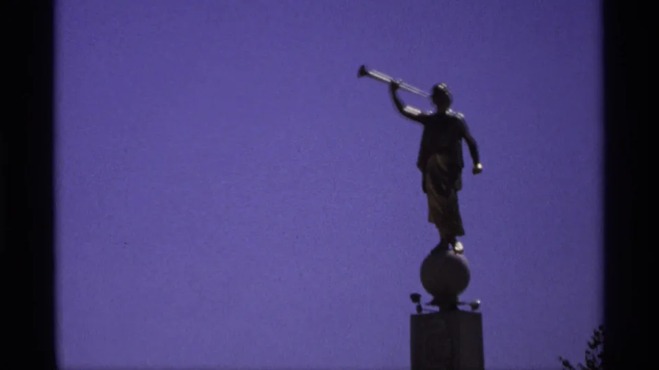 Dark horn-blowing statue on a pedestal, silhouetted against a purple-blue sky in 1970s 8mm footage.