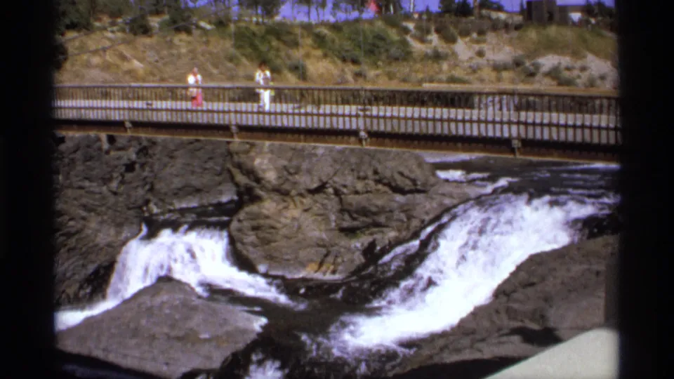 Distant people cross a pedestrian bridge over white rushing water and dark rocks in archival 8mm footage.