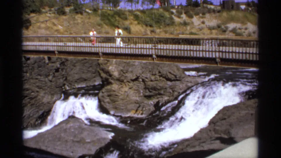 A rocky waterfall flows beneath a railing-lined pedestrian bridge where visitors walk in bright daylight.