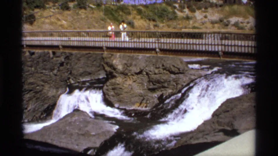 People stand and walk along a bridge spanning a rocky waterfall area, with hillside landscaping behind them.
