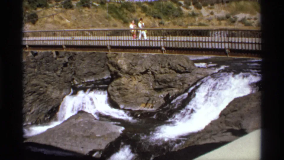 The camera frames whitewater moving through dark rocks below a public pedestrian bridge with visitors above.