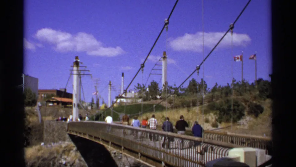 Visitors walk across a cable-supported pedestrian bridge near flags and landscaped slopes under a blue sky.