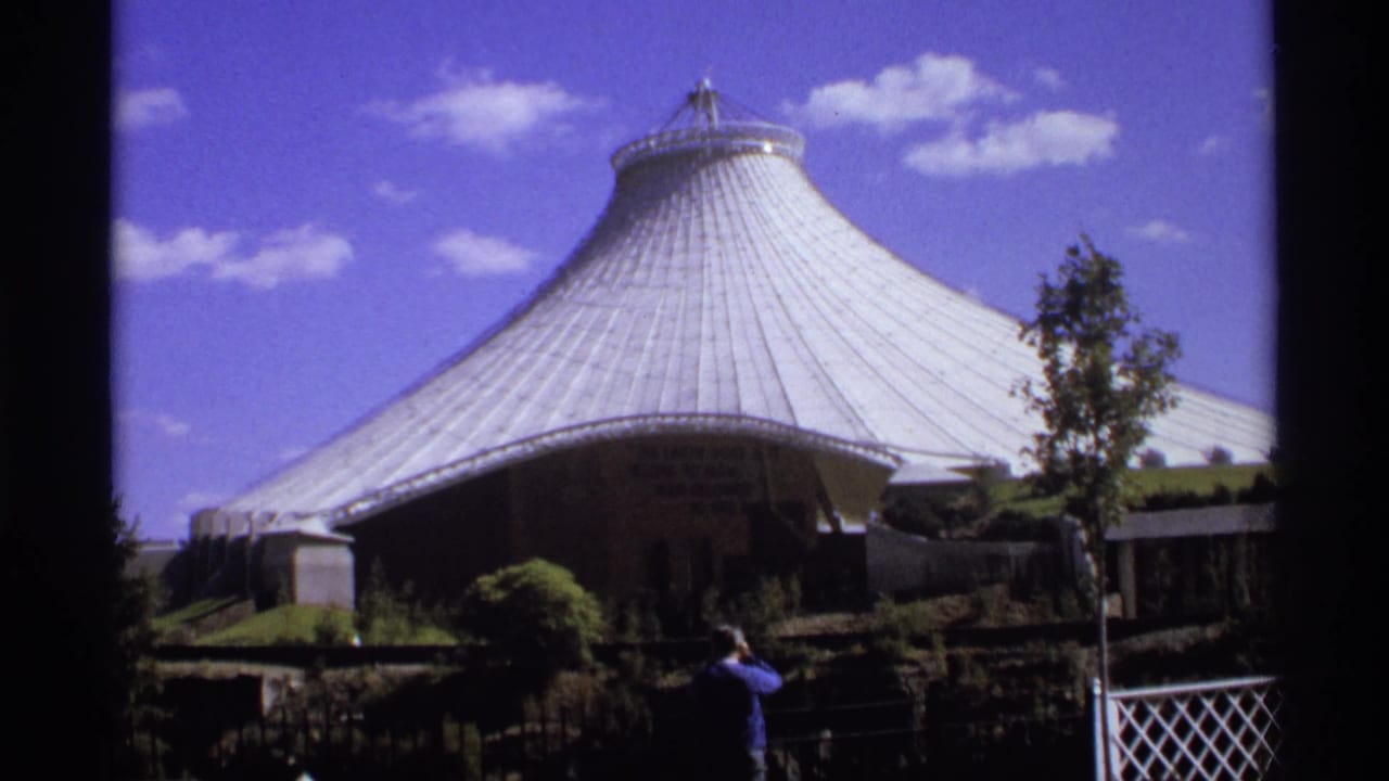 1974 8mm frame showing a large ribbed dome, landscaping, blue sky, and a visitor facing the structure.