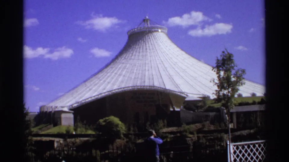 1974 8mm frame showing a large ribbed dome, landscaping, blue sky, and a visitor facing the structure.