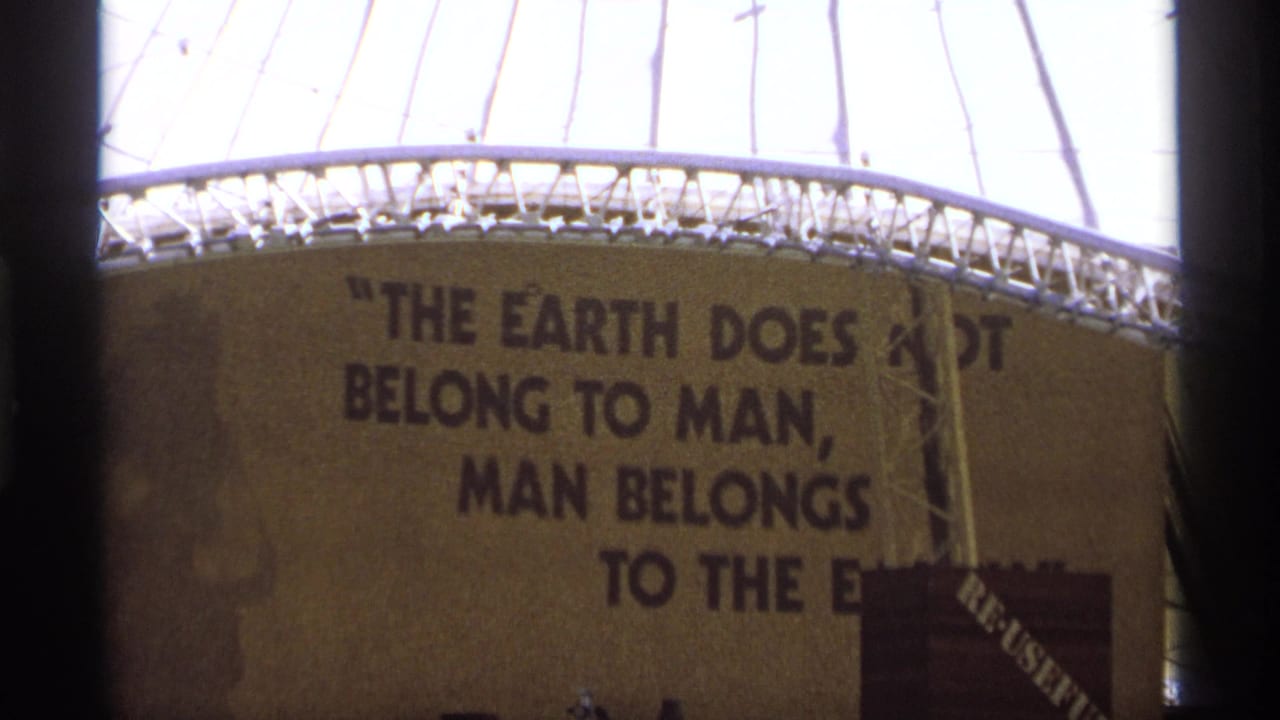 1970s 8mm frame of an exhibit-like wall with a large environmental quote, partially obscured by dark vertical edges.