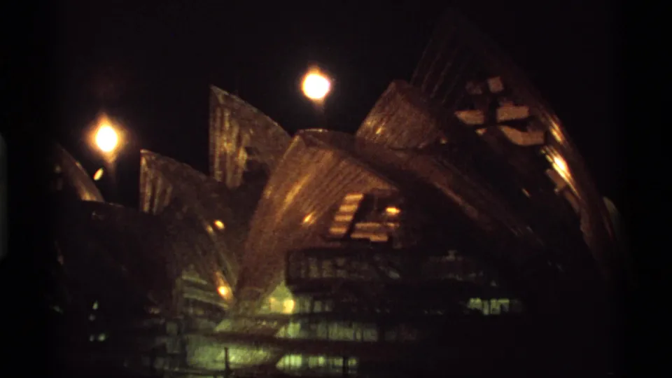 Night 8mm frame of a warmly lit shell-like architectural structure, resembling the Sydney Opera House, seen from a visitor viewpoint.