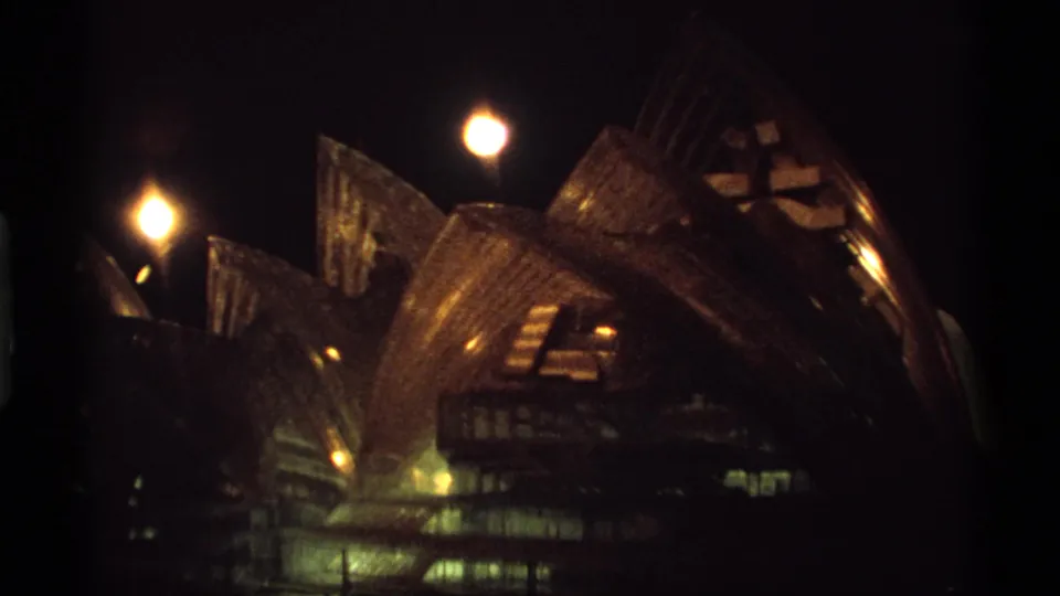 Handheld 8mm night view of illuminated shell-like architecture with warm lamps and dark framing at the edge.