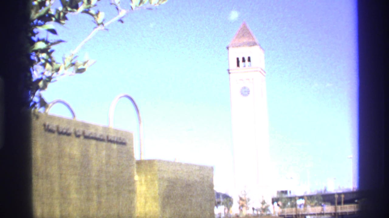 White clock tower beside a stone building with arched metal elements under bright blue sky.