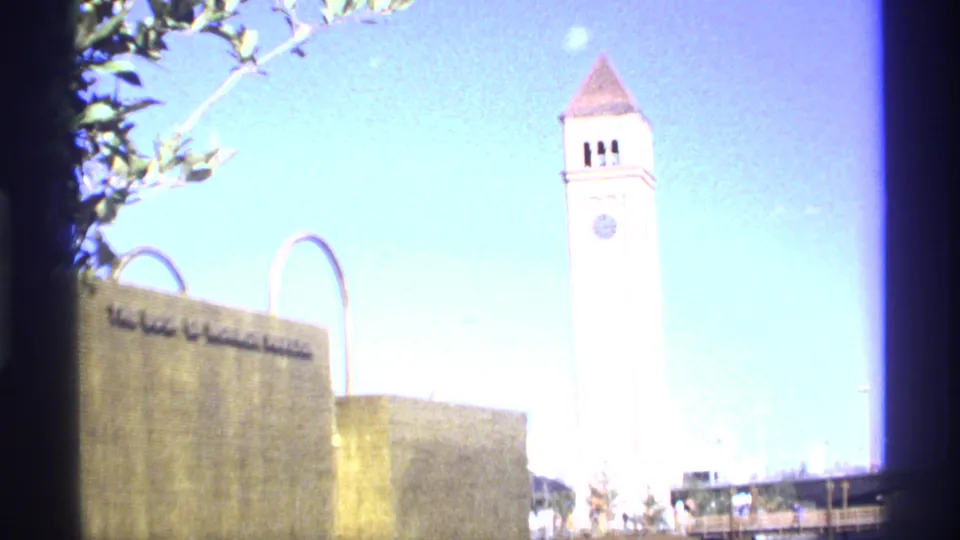 Daylight 8mm frame showing a tall white clock tower, a stone building with partial lettering, metal arches, blue sky, and tree branches.