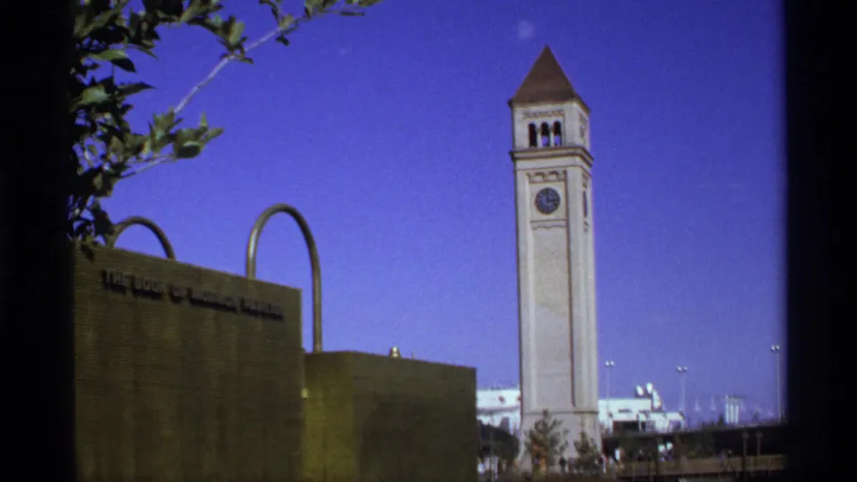 Handheld 8mm daylight frame with a tall white clock tower, stone wall with partial text, arched metal forms, tree branches, and distant vehicles.