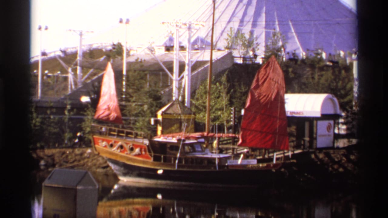 Red-sailed Chinese junk-style boat on calm water with a geodesic dome behind it.